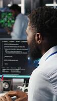 Vertical Server farm computer scientist interprets data analytics graphs. African american man in data center using artificial intelligence program showing predictive system metrics, camera A video