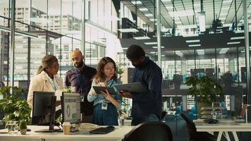 Multiethnic coworkers looking over revenue figures on tablet in diverse office. Multiracial colleagues doing brainstorming, comparing company statistical numbers, working hard to finish job, camera B video