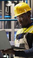 Vertical Professional working in smart factory, testing automated systems to ensure reliability. African american man in industry 4.0 plant using notebook to identify bottlenecks and optimize, camera A video