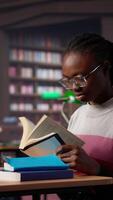 vertical Preto menina focos em dela exame preparativos dentro a campus biblioteca, leva notas em dela computador portátil a partir de referência livros dentro uma pacífico estude contexto. menina usa a geral base de dados materiais. Câmera uma. video
