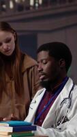 Vertical Diverse group of pupils in a library discuss with a medic resident, preparing for a new lecture at med school. Using medical books and materials to gain expertise in microbiology. Camera A. video