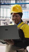 Vertical Smiling smart factory engineer typing on laptop, overseeing monitoring of production lines. Joyful woman in industry 4.0 plant ensures machinery operates at optimal efficiency using notebook, camera A video