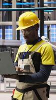 Vertical Portrait of smiling technician in solar panels factory oversees robot assembly lines, ensuring quality control. African american man in industrial plant monitoring automated conveyor systems, camera A video
