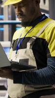 Vertical Smart solar panels factory technician using laptop to oversee conveyor belts. African american man in industry 4.0 plant ensuring machinery operates at optimal efficiency using notebook, camera B video