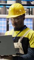 Vertical Portrait of smiling technician in solar panels factory oversees robot assembly lines, ensuring quality control. African american man in industrial plant monitoring automated conveyor systems, camera B video