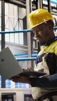 Vertical Smart solar panels factory technician using laptop to oversee conveyor belts. African american man in industry 4.0 plant ensuring machinery operates at optimal efficiency using notebook, camera A video
