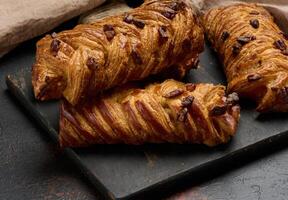 Three golden brown maple pecan braid pastries are arranged on a dark, textured surface. Braided dough is flaky and topped with pecans. photo