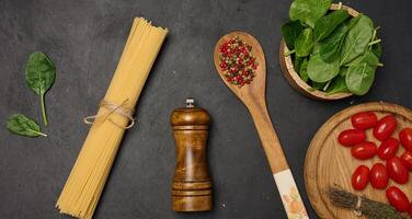 Fresh spinach, cherry tomatoes, spaghetti, and spices on a dark rustic surface. A wooden cutting board, thyme bundle, and peppercorns, top view photo
