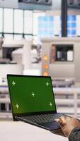 Vertical Factory engineer typing on green screen laptop, monitoring production lines. Woman in industry 4.0 plant ensures machinery operates at optimal efficiency using chroma key mockup notebook, camera A video