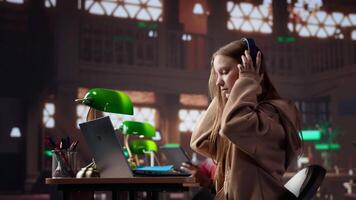 Young girl listening to educational documentary on laptop and takes notes, using modern non conventional way to study in the university library. Female student collects data. Camera A. video