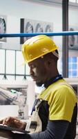 Vertical Technician in smart factory using notebook to look at system performance metrics. African american man in industry 4.0 plant using laptop to track energy production through IoT sensors, camera A video