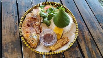 A delicious portion of fried tilapia served with white rice and traditional sambal on a woven bamboo tray . A classic Indonesian dish presented in a rustic, cultural style. photo