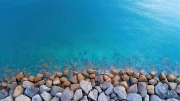 Aerial view of turquoise water with rocky shoreline and visible underwater stones along a calm coastal edge under sunlight. photo