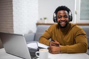 A young black freelancer working from home, using his laptop to communicate with clients and taking notes during a conference. photo