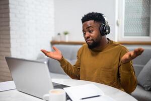 Confused young man with headphones gesturing and talking while attending e-learning session via laptop from the comfort of his home. photo