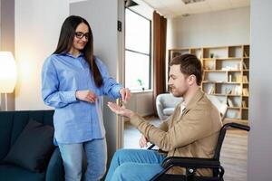 Man in wheelchair extend hand palm up to side. Woman giving key with keychain from new house for handicapped. photo
