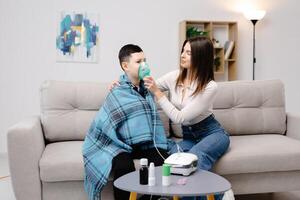 Young woman helping illness boy with nebulizer inhalation at home. A young mother helping her son to inhale with a nebulizer at home. photo