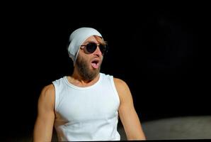 Man wearing a tank top, sunglasses, and a beanie poses confidently at night against a dark background. photo