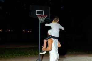A father and son share a fun moment playing basketball outdoors at night with a hoop in the background. photo