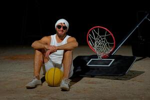 Basketball player sits on the court wearing sunglasses, relaxed after an intense practice session at dusk. photo