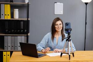 Students learning remotely online using web app. Female teacher providing lesson with laptop, recording on cellphone on tripod photo