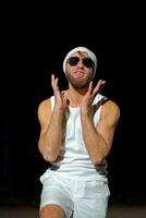 Man wearing a tank top, sunglasses, and a beanie poses confidently at night against a dark background photo