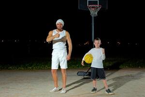 Smiling adult parent with cute child coming at outdoor court to play basketball. Father and son with ball talking while walking. Playground in city park. photo