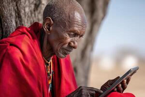 Elderly man in Maasai attire uses tablet under a tree in the African savannah photo