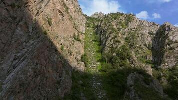 Aerial footage showing a forward approach to a rugged mountain chute in Kyrgyzstan, revealing a natural rocky path winding through steep cliffs and sparse greenery under a clear blue sky. video