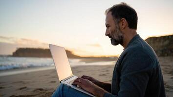 A man using a laptop on a beach at sunset, representing freedom and remote work. photo