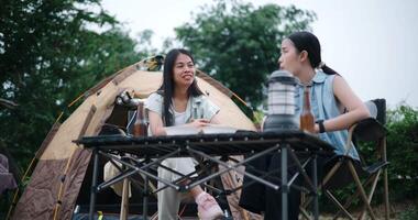 Handheld low angle view shot, Two Asian women sit together in a garden, happily chatting while enjoying snacks and drinks, surrounded by a cozy camping vibe. Holiday Activities video