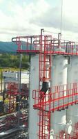 Aerial view gas station operator descends from the top of the station. Modern gas complex in the mountains. A gas worker climbs the ladder of the gas distribution unit, against the backdrop of video
