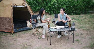 Handheld high angle view shot, Two Asian friends sit on chairs and talk happily in front of a tent in a camping atmosphere. video