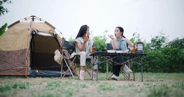 Handheld low angle view shot, Asian women sit together in a garden, happily chatting while enjoying snacks and drinks, surrounded by a cozy camping vibe. Holiday Activities video