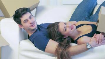In a cozy, empty apartment, a loving couple lies together on a white couch, surrounded by cardboard boxes, symbolizing their new beginning. Their smiles radiate joy and excitement as they embrace, ready to unpack their dreams in this fresh space video