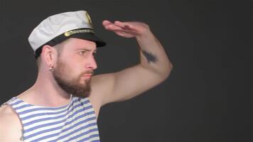 A stylish young bearded man in a striped sailor singlet salutes with his hand on his forehead, exuding confidence and charm. This Caucasian hipster, adorned with a nautical cap, stands against a sleek dark gray background, embodying a modern maritime vibe video