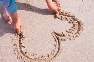 Child drawing heart shape in sand on beach photo