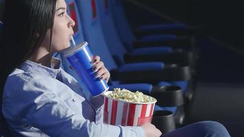 un asiático niña se sienta en perfil, bebiendo un refrescante soda y participación un grande Cubeta de palomitas de maiz en un vibrante cine. rodeado por amigos, el atmósfera es lleno con emoción y alegría como ellos sumergirse sí mismos en el cinematográfico experiencia video