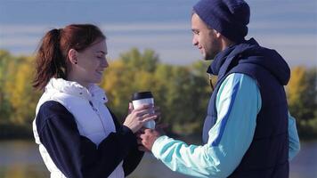 A cheerful young couple enjoys a moment together, with the man handing a warm coffee cup to his girlfriend against a stunning autumn backdrop. Their smiles radiate joy and connection, embodying the spirit of fitness and love on a crisp morning video