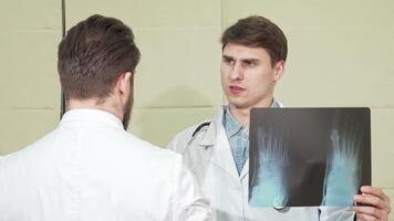 A focused male doctor, with a stethoscope around his neck, engages in a detailed discussion about a foot x-ray with his colleague. The bright clinic setting enhances the atmosphere of collaboration and expertise in the medical field video