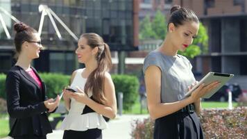 In a vibrant urban setting, a poised young businesswoman reads the news on her tablet, exuding professionalism. Meanwhile, her stylish colleagues share a light moment, showcasing a dynamic atmosphere of collaboration and success video