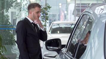 A young businessman in a sharp suit stands pensively, examining a stylish car at a modern dealership. His thoughtful expression and the gleaming vehicles around him create an atmosphere of excitement and anticipation for his next big purchase video