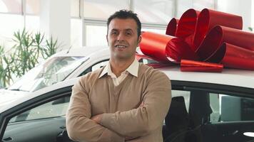 A solidly built man with dark hair stands confidently, arms crossed, in front of a sleek car topped with a vibrant red gift bow. His warm smile radiates excitement, inviting viewers to share in the joy of this perfect new purchase video