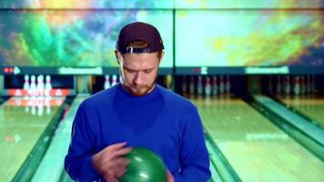 A young Caucasian man in a cap confidently gestures 'OK' while holding a vibrant green bowling ball, ready to take his shot. The lively bowling alley backdrop, with colorful lights and neatly arranged pins, sets an energetic scene for this fun-filled game video