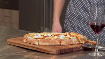 A young man joyfully reaches for a slice of hot, delicious pizza, perfectly placed on a rustic wooden board. The inviting aroma fills the cozy apartment, complemented by a glass of rich red wine, creating a delightful evening of relaxation and indulgence video