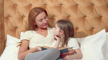An engaging mother and her adorable little daughter share a cozy reading moment on a plush bed, surrounded by soft pillows. This heartwarming scene captures the joy of home schooling, where imagination and knowledge intertwine in a nurturing environment video