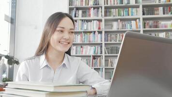 A joyful Asian woman beams with laughter as she engages with her laptop in a cozy library setting. Surrounded by shelves of colorful books, she enjoys a light-hearted moment, taking a break from her studies and immersing herself in entertaining content video