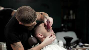 In a chic barbershop, a skilled barber meticulously shaves a client's beard, showcasing precision and artistry. The warm ambiance, highlighted by vintage decor and soft lighting, enhances the grooming experience, making it a moment of relaxation and style video