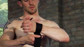 A focused shirtless male MMA fighter with a chiseled physique meticulously wraps his hands in preparation for an intense bout. The gritty gym backdrop enhances the atmosphere of determination and strength, showcasing the athlete's commitment to victory video
