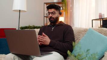 Indian man using wireless headset speaks on laptop call with client while working at home video